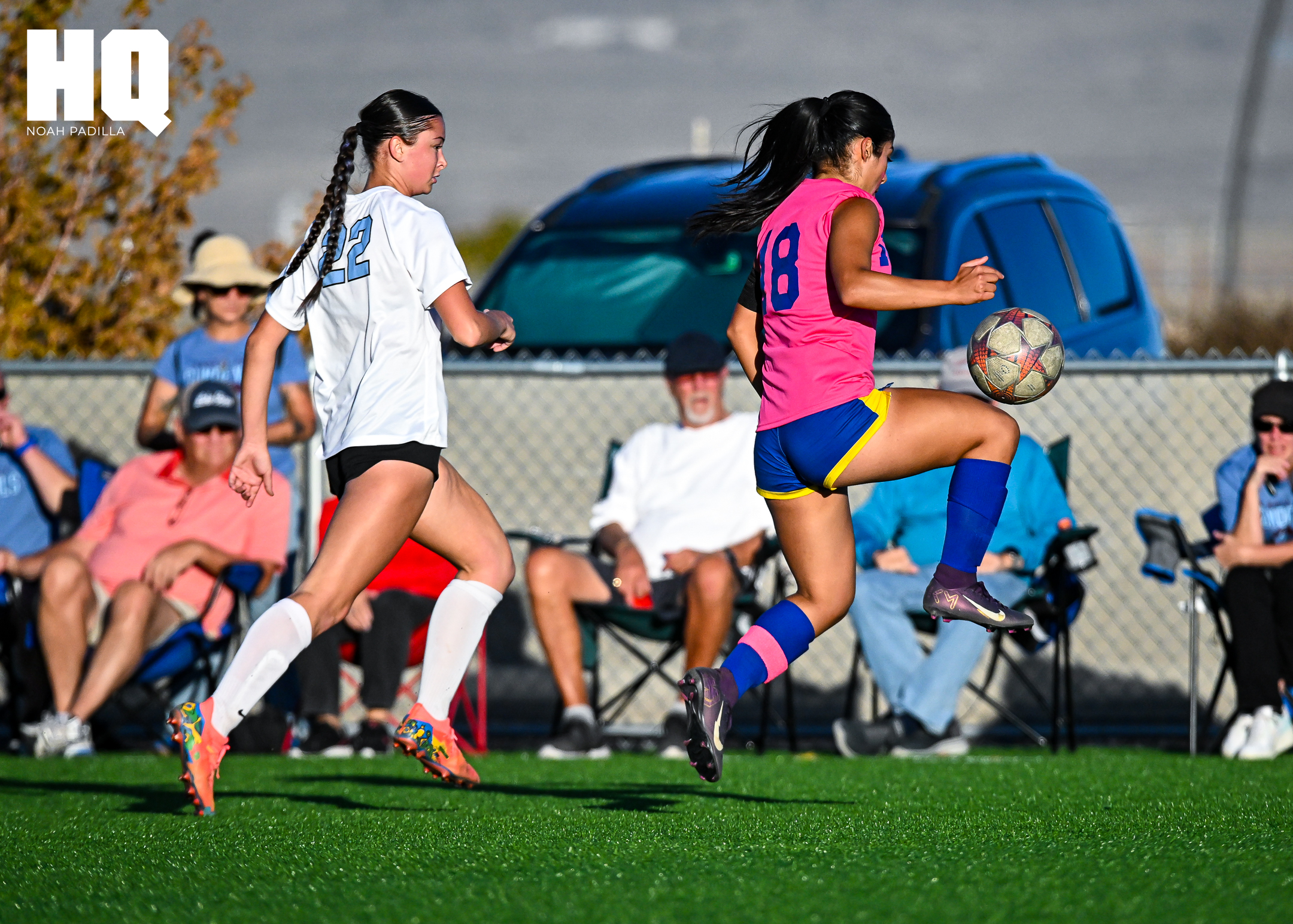 A Cottonwood Classical player, Savannah Serbin, in a pink jersey with number 18 controls the ball in midair with her knee as a Sandia Prep defender wearing number 22 trails behind during live play.