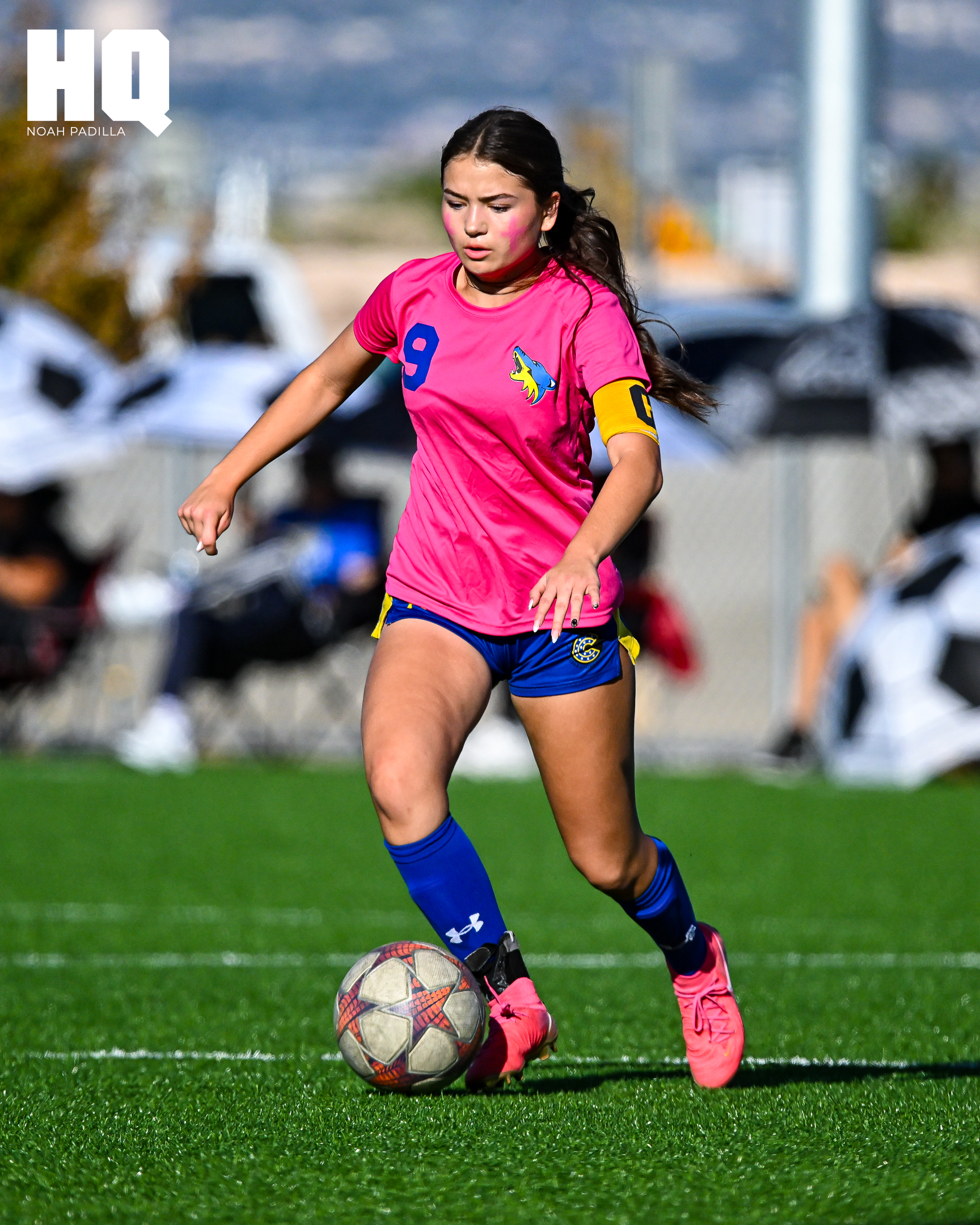 Sophia Soriano of Cottonwood Classical girls soccer wearing a pink jersey with number 9 and a captain’s armband dribbles the ball upfield with intensity during a match on a turf field.