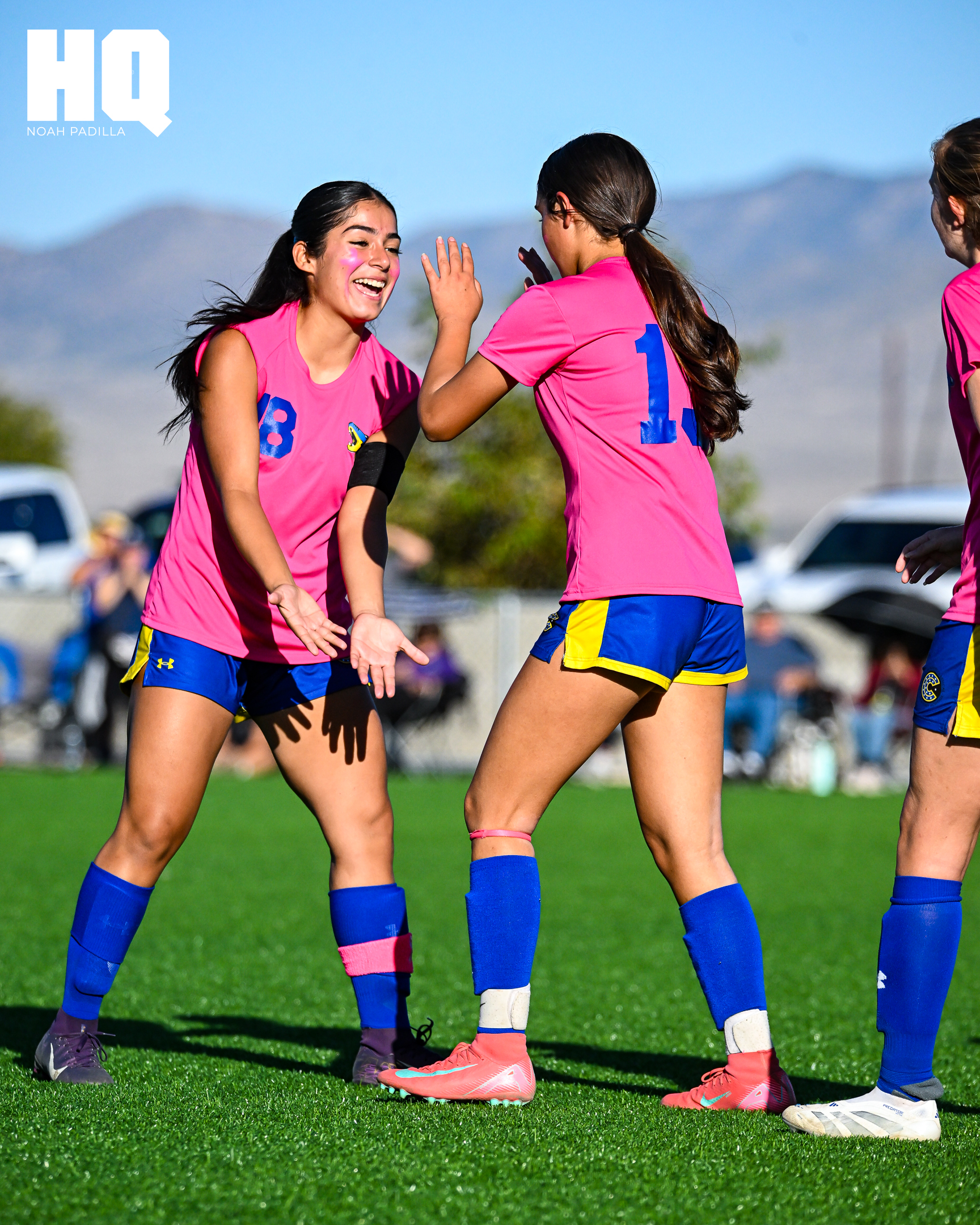 Cottonwood Classical players, #18 Savanah Serbin and #13 Hailo Baca, in pink jerseys celebrate with a high-five and smiles, showcasing team spirit and joy after Baca's goal. A third teammate watches with a smile in the background.